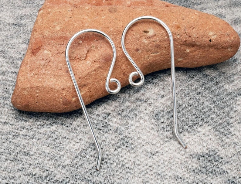 K&ouml;nnte beinhalten: Zwei silberfarbene Ohrhaken. Jeder Haken hat eine gebogene Oberseite mit einer kleinen, aufgerollten Schlaufe und einen geraden, schlanken Draht, der sich am Ende kr&uuml;mmt. Die Haken werden auf einem strukturierten, rotbraunen Stein vor grauem Hintergrund gezeigt.