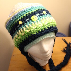 May include: A crocheted hat with a blue, green, and white color pattern. The hat has a long, braided blue scarf attached to it. There are three yellow buttons on the hat.
