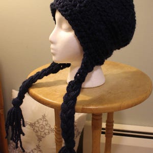 May include: A navy blue crocheted hat with long, braided tassels. The hat is displayed on a mannequin head, showcasing its shape and design. The tassels hang down, adding a playful element to the winter accessory.