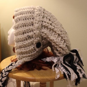 May include: A handmade, light gray crochet hat with a black button detail. The hat has a braided black and white accent and a decorative fringe of black and white lace. The hat is displayed on a mannequin head.