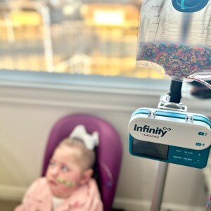 May include: A young girl sits in a purple chair, looking to the right. A medical device with the text "Infinity - MOOG" is attached to a clear bag filled with colorful sprinkles.