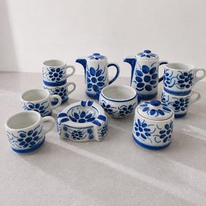 May include: A ceramic tea set featuring a white base with hand-painted blue floral patterns. The set includes two teapots with lids, a lidded sugar bowl, an open bowl, a triangular ashtray, and five mugs, all decorated with matching blue motifs.