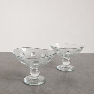 Puede incluir: Dos cuencos de postre de vidrio transparente con una superficie texturizada y una serie de puntos redondos en relieve. Cada cuenco tiene una base redondeada y un tallo. Los cuencos están sobre una superficie gris.