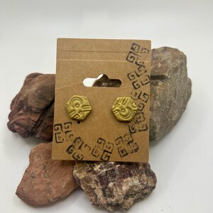 Könnte beinhalten: Ein Paar goldfarbene Ohrringe mit geometrischem Design. Die Ohrringe werden auf einer braunen Karte mit einem schwarzen Stempel präsentiert.