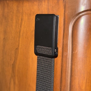 May include: A black belt buckle with a textured nylon strap. The buckle is rectangular with a small, subtle design on the front. The strap is a dark gray color and appears to be made of a durable material.