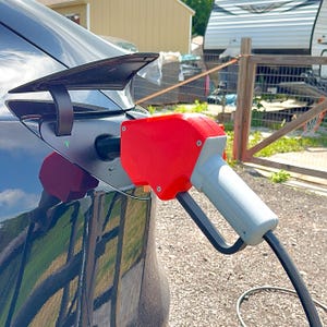 Op de afbeelding: Een rode en grijze elektrische voertuig oplaadconnector aangesloten op een zwarte auto. De oplaadpoort is open en de connector zit stevig vast. De auto staat buiten op een zonnige dag.