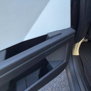 Puede incluir: Primer plano del interior de una puerta de coche. La puerta presenta un reposabrazos negro con una pequeña manija rectangular gris. Debajo del reposabrazos hay un compartimento de almacenamiento. El panel de la puerta es blanco. La puerta del coche está abierta.
