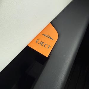 May include: Close-up of an orange car eject button with the word "EJECT" in white text. The button has a graphic of a car with an arrow above it. The button is located between a white and black surface.