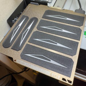 Op de afbeelding: Een goudkleurig, getextureerd oppervlak met verschillende donkergrijze rechthoekige stukken. Elk stuk heeft een wit geometrisch ontwerp. De stukken zijn op het oppervlak gerangschikt, wat wijst op een mogelijke lay-out of patroon.