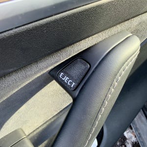 May include: Close-up of a black car door handle with the word "EJECT" printed in white. The handle is made of black leather and has white stitching. The surrounding area is dark gray.