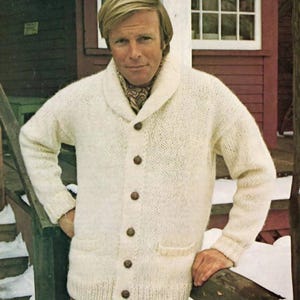 May include: Cream-colored, chunky knit cardigan with a shawl collar and wooden buttons. The sweater has two front pockets and is worn with brown corduroy pants. The person is standing outside near a wooden railing.