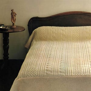 May include: A cream-colored, patterned knitted blanket covers a bed with a dark wooden headboard. The blanket features various textured designs, including vertical stripes and geometric patterns. A small, round side table with a statue is visible.