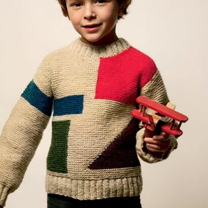 Op de afbeelding: Een kind draagt een beige gebreide trui met een kleurblokontwerp in rood, groen, blauw en bruin. Het kind houdt een klein rood houten vliegtuig vast. De trui heeft een geribbelde halslijn, manchetten en zoom.
