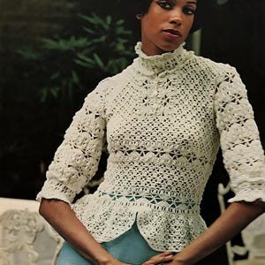 May include: A cream-coloured, open-weave crochet top with a high collar and three-quarter sleeves. The top has a fitted bodice and a flared peplum. The model is wearing a light blue skirt. The image has a vintage aesthetic.