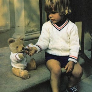 Puede incluir: Un niño con un suéter blanco, pantalones cortos azul marino y zapatos negros está sentado junto a un oso de peluche. El oso de peluche también lleva un suéter blanco. El niño le ofrece algo al oso.
