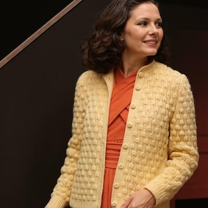 Peut inclure: Un cardigan jaune clair avec un motif à bulles texturé. Le cardigan a une fermeture boutonnée sur le devant et des manches longues. La personne porte une robe orange en dessous. Le fond est un mur sombre.