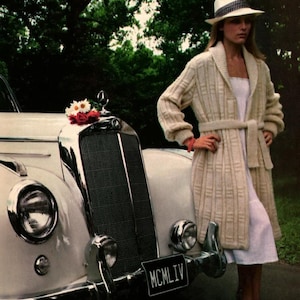 May include: A woman in a cream-coloured, long knit cardigan and a white dress stands beside a vintage white car. She wears a white hat with a dark band. The car has a chrome grill and headlights, with a license plate reading "MCMLIV".