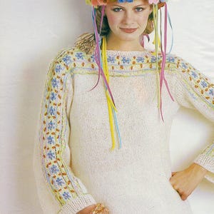 May include: A cream-coloured sweater with floral embroidery on the sleeves and neckline. The sweater is paired with a flower crown and colourful ribbons. A floral bracelet is also visible. The overall aesthetic is bohemian and vintage.
