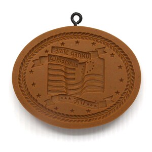 May include: A brown, oval-shaped cookie mold with a raised design of the American flag and the words "UNITED AMERICA" and "FLAG". The mold has a decorative rope border and a small metal loop for hanging.