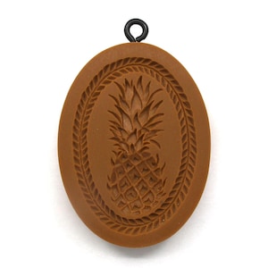May include: An oval-shaped brown pendant featuring a debossed pineapple design in the center, surrounded by a textured rope-like border. A small black metal jump ring is attached to the top of the pendant for hanging or connecting to a chain.