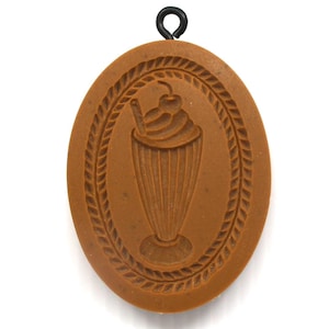 May include: An oval-shaped brown cookie mold featuring a debossed illustration of a milkshake topped with whipped cream and a cherry, surrounded by a decorative rope-style border. A small black metal loop is attached to the top for hanging.