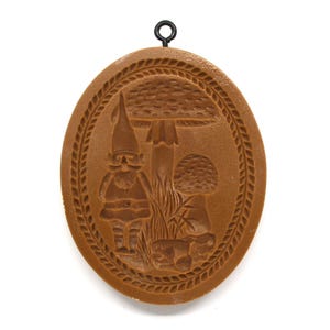 May include: Brown oval-shaped mold with a gnome and mushroom design. The gnome wears a pointed hat and has a long beard. The mold has a decorative border and a metal loop for hanging.