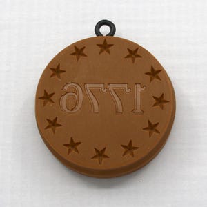 May include: A circular brown silicone mold featuring the embossed text 1776 in reverse, surrounded by a ring of thirteen five-pointed stars. A small black circular loop is attached to the top edge of the mold for hanging or attachment purposes.