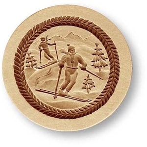 May include: A round, light brown wooden carving featuring two skiers, trees, and mountains. The skiers are in vintage ski suits, and the design is framed by a dark brown, textured border. The overall theme is winter sports.