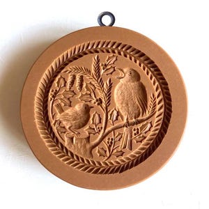 May include: A brown cookie cutter with a raised design of two birds perched on a holly branch. The cutter has a metal loop at the top for hanging.
