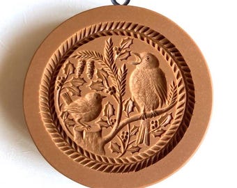May include: A brown cookie cutter with a raised design of two birds perched on a holly branch. The cutter has a metal loop at the top for hanging.