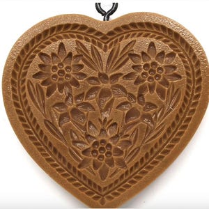 May include: A heart-shaped brown mold featuring an embossed design of three edelweiss flowers surrounded by a braided border. The mold has a small black metal loop at the top center for hanging, with a textured surface throughout the piece.
