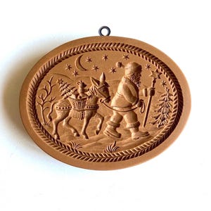 May include: A brown wooden cookie cutter shaped like a Christmas scene with Santa Claus walking with a donkey carrying a Christmas tree and presents. The cookie cutter has a rope border and a hanging loop.