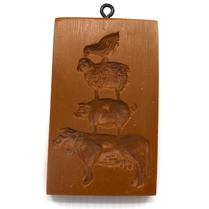 May include: A rectangular, brown, wooden mold featuring a cow, pig, sheep, and chicken stacked on top of each other. The mold has a black metal loop at the top for hanging. The farm animal design is carved into the wood.