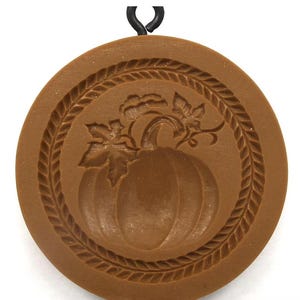 May include: A round, brown, flat-surfaced mold featuring a central embossed pumpkin design with leaves and a vine, surrounded by a decorative braided border. A small black metal loop is attached to the top edge for hanging or display purposes.