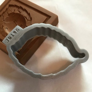 May include: A gray plastic cookie cutter shaped like a leaf with a textured edge. The cutter is sitting on a brown surface with the number "3019B" printed on it.