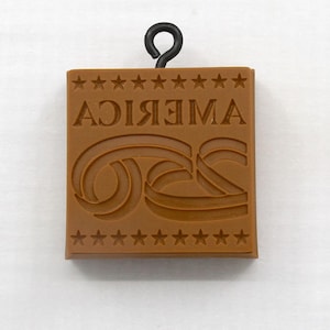 May include: A brown, square-shaped pendant with the word "AMERICA" in reverse and a stylized "GS" design. The pendant is adorned with star accents and has a black metal loop at the top for hanging.