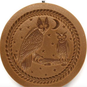 May include: A circular brown cookie mold featuring a relief design of two owls perched on a branch under a crescent moon and stars. The central image is enclosed within a decorative braided border, creating a textured surface for baking.