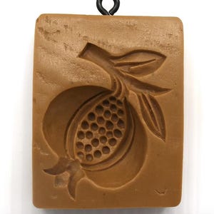 May include: A rectangular light brown mold featuring a carved pomegranate design with a stem and leaves. The pomegranate fruit is depicted with a textured interior showing seeds. A small black metal loop is attached to the top edge of the mold.