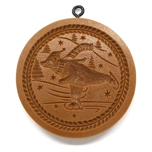 May include: A brown, circular cookie mold with a detailed relief of a bear ice skating. The design includes trees, stars, and a decorative border. A black metal loop is attached at the top for hanging.