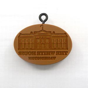 May include: A brown oval-shaped stamp featuring an embossed image of The White House. The text "THE WHITE HOUSE WASHINGTON" is also embossed on the stamp. A black metal loop is attached at the top.