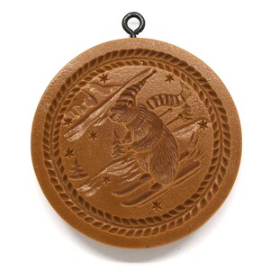 May include: A round, brown, decorative ornament with a black metal loop at the top. The ornament features a skier with skis and poles, surrounded by stars and a braided border. The skier is wearing a hat and scarf.