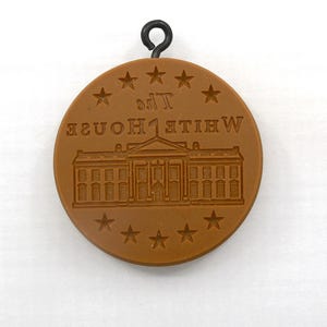 May include: A brown, circular pendant featuring an embossed image of the White House and the text "THE WHITE HOUSE" in reverse. The pendant has a black metal loop at the top and is surrounded by embossed stars.
