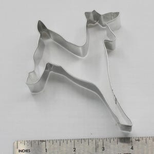May include: A silver-colored metal cookie cutter in the shape of a reindeer. The reindeer is posed in a running position. The cutter is approximately 4 inches long, as measured by the ruler in the image. The cutter is on a white surface.