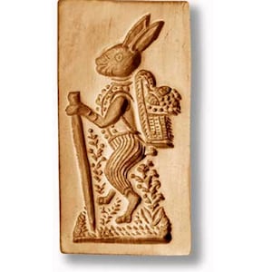 May include: A wooden cookie cutter shaped like a rabbit carrying a basket on its back. The rabbit is wearing a vest and pants and is holding a stick in its hand.