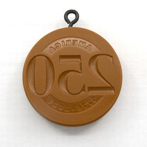 May include: A round, brown pendant with the embossed text "250 AMERICA" and "SOS-OTTO" in a circular pattern. A black metal loop is attached at the top for hanging. The pendant has a vintage or antique appearance.