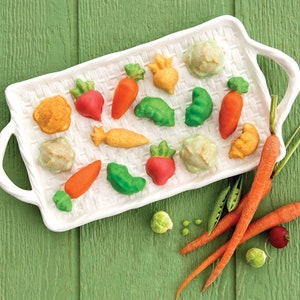 May include: A white rectangular serving tray filled with colorful, miniature vegetable-shaped candies. The candies include carrots, radishes, peas, and cabbage. Fresh carrots, peas, and a brussel sprout are arranged on a green wooden surface.