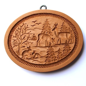 May include: A wooden cookie cutter shaped like a winter scene with deer, rabbits, and pine trees. The cutter has a brown finish and a hanging loop.