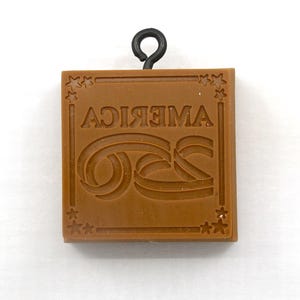 May include: A brown, square-shaped stamp with the word "AMERICA" in reverse and a stylized "GS" design. The stamp has a black wire loop at the top for hanging and star accents in each corner. The stamp is likely used for creating impressions.