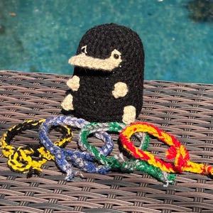 May include: A handmade black crochet Niffler with a tan snout and paws, sitting above several colorful braided bracelets. The bracelets are yellow, blue, green, and red, and are made of yarn.