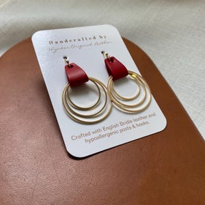May include: A pair of gold hoop earrings with three rings each. The hoops are attached to red leather tabs. The earrings are displayed on a white card with the text "Handerafted by Wynker Original Leather" and "Crafted with English Bridle leather and hypoallergenic posts & hooks."